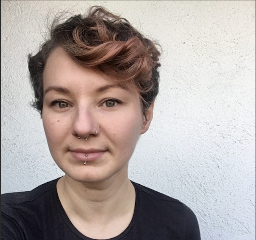 a headshot of a white young adult with a reddish hair in a pixie cut wearing a black shirt. the person smiles slightly as they look into the camera at a slight leftward angle against a gray-white background.